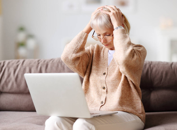 Erschöpfte reife Frau sitzt mit Laptop auf Sofa und berührt Kopf, während sie unter Müdigkeit und Kopfschmerzen leidet - Foto, Bild