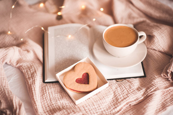 Cup of coffee staying on open book with heart shape cookie over lights close up. Good morning. Breakfast time. Valentines Day. - Photo, Image