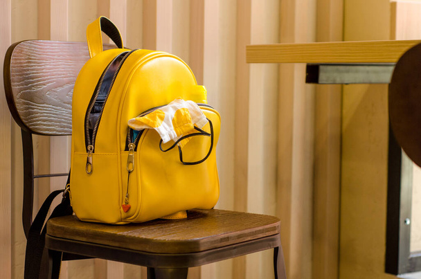 Yellow urban backpack with a white protective mask in his pocket, stands on a wooden chair in a cafe near table and striped wall. - Photo, Image