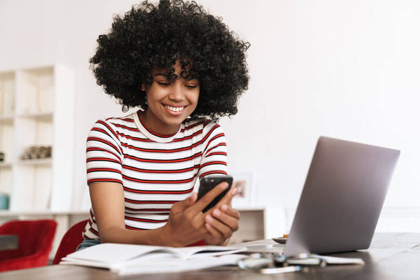 Smiling african american student girl using cellphone while studying with laptop in cafe - Фото, зображення