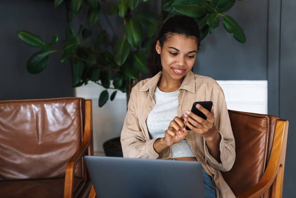 Usmívající se africká americká žena pracující s notebookem a mobilním telefonem, zatímco sedí v kavárně - Fotografie, Obrázek