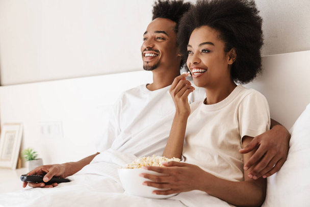Couple sitting in bed together eating popcorn, holding television remote - Valokuva, kuva