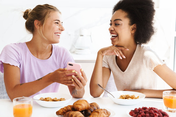 Nice cheerful multicultural girls using cellphone while having breakfast together at home kitchen - Φωτογραφία, εικόνα