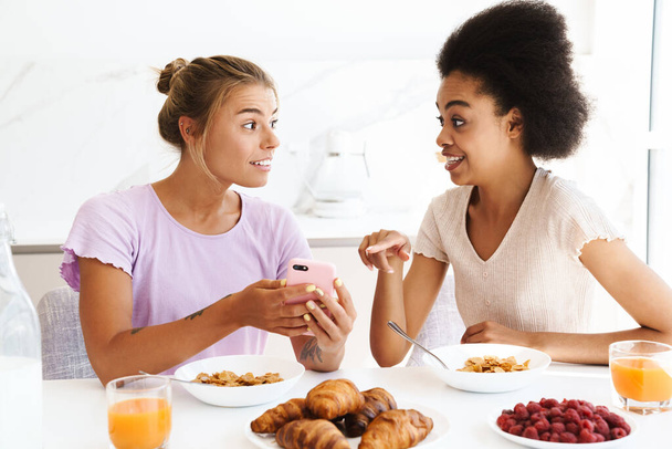 Nice surprised multicultural girls using cellphone while having breakfast together at home kitchen - Фото, зображення