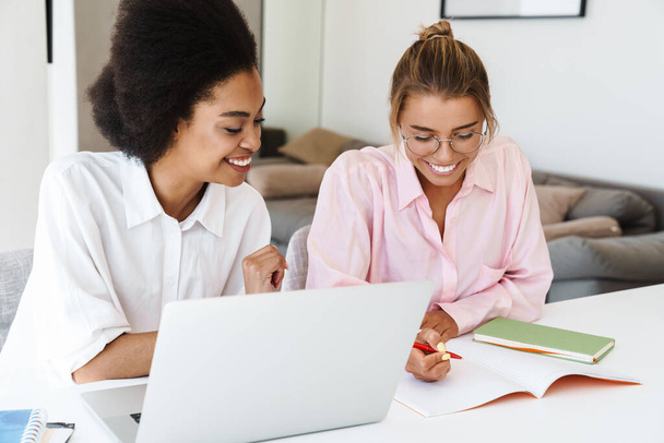 Joyful multicultural students girls doing homework with laptop together at home - Фото, изображение