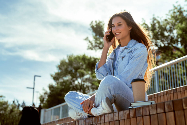 Millennial girl in denim clothes sits on park bench, communicates with friends, colleagues, parents on the phone. - Фото, изображение