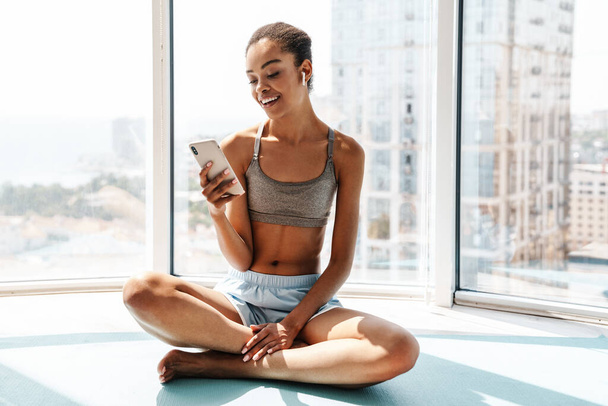 Image of a young cheerful sports african woman using mobile phone and listening music with earphones indoors - Photo, image