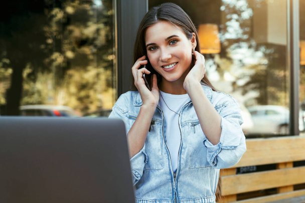 Jovem mulher de negócios trabalhando no terraço aberto em casa, sentada na frente do laptop, segurando smartphone em sua mão.  - Foto, Imagem