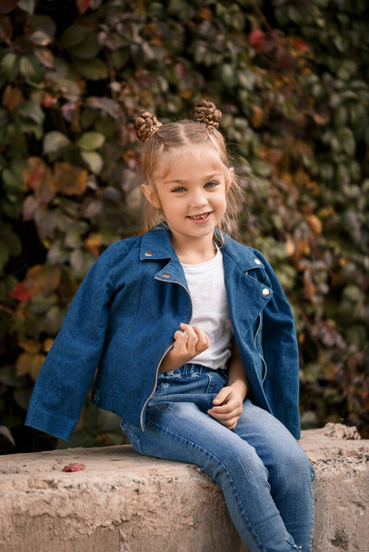 Una bella ragazza bionda elegante in una giacca blu si siede su un blocco di cemento. Ragazza di 7 anni con un sorriso su uno sfondo di foglie - Foto, immagini