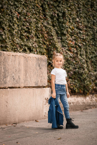 Stylowa blondynka w dżinsach i biała koszulka spacerują ulicą. Dziewczyna 7 lat mały model, piękne dziecko - Zdjęcie, obraz