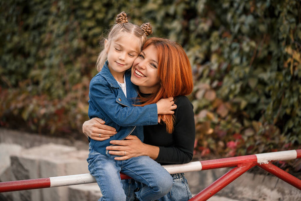 スタイリッシュな赤い髪の母親と彼女の娘は暖かく話しています。路上で幸せな子供と彼女の母親。散歩のための彼女の娘との成功したシングルマザー。暖かい家族関係 - 写真・画像