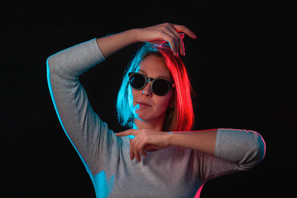 The neon lights of the club. Portrait of a young woman in glasses, posing with her hands. Side blue and red light. Black background. - Фото, зображення
