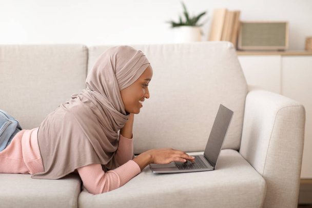 Side view of black woman in hijab using laptop with blank screen on sofa at home, copy space - Foto, Imagem