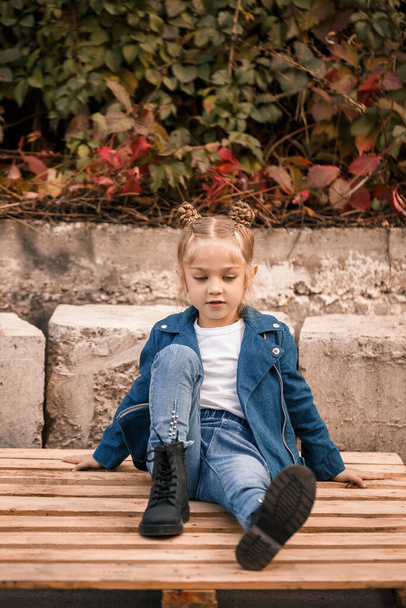 Une belle petite fille est assise dans des vêtements élégants sur des palettes en bois. Enfant élégant dans une veste bleue. Style décontracté, mode pour enfants, costume à la mode, enfance heureuse - Photo, image