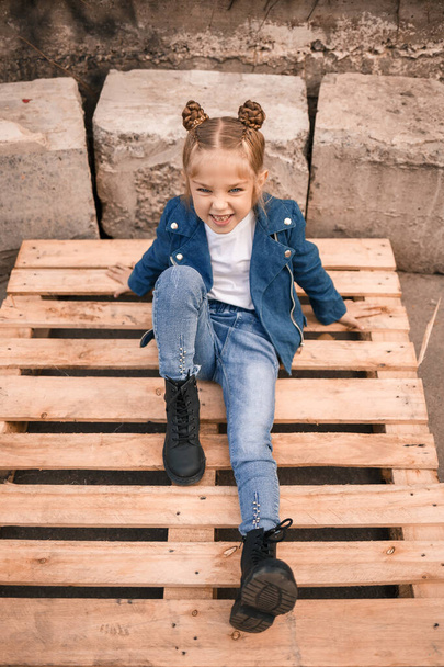 Una hermosa niña está sentada en ropa elegante en paletas de madera. Niño con estilo en una chaqueta azul. Estilo casual, moda para niños, traje de moda, infancia feliz - Foto, Imagen