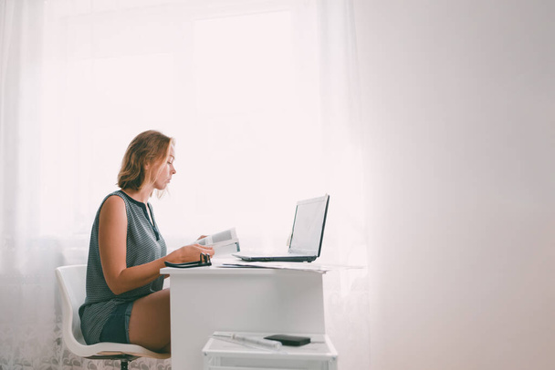 Een jonge vrouw zit aan een tafel en bladert door een boek. Vlakbij zijn een laptop en kantoorbenodigdheden. Het interieur is wit van kleur. Kopieerruimte. - Foto, afbeelding