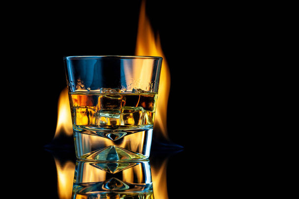 Whiskey or bourbon in a transparent glass with ice cubes on a black background with fire. Horizontal template for design. Place for your text. - Foto, imagen