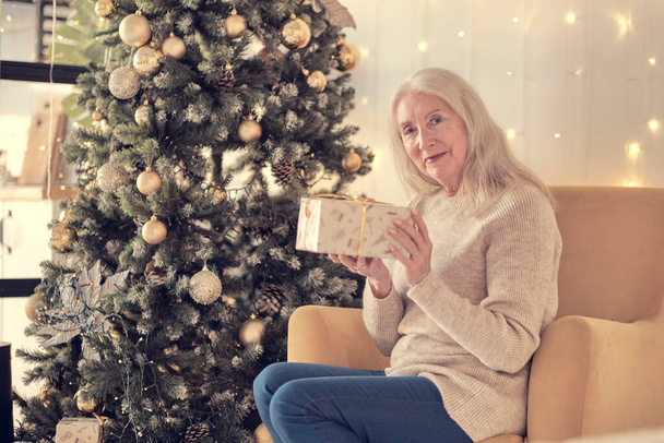 La anciana positiva está descansando en el sofá y mostrando regalo de Navidad - Foto, Imagen