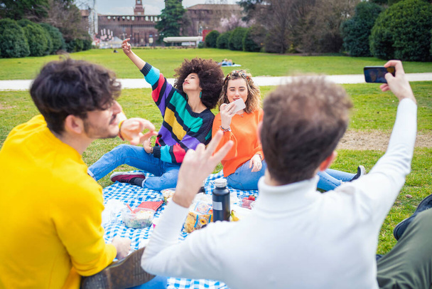 Four young students multi ethnic friends outdoor doing pic nic in a park spending time together and having fun celebrating happy moments doing selfie and photos collecting memories - Photo, Image