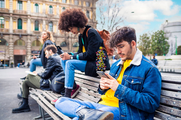 Τέσσερις πολυεθνικοί άνθρωποι ξένοι ή φίλοι κάθονται στον πάγκο υπαίθρια χρησιμοποιώντας smartphone αγνοώντας εθισμένοι στις κοινωνικές τάσεις μετά τη χρήση emoticons - Φωτογραφία, εικόνα