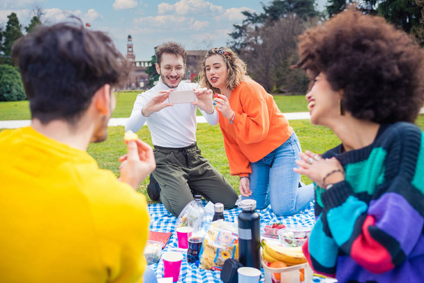 Quatre amis joyeux divers multiethnique s'amusent à faire un pique-nique dans un parc à l'extérieur en utilisant un smartphone prenant des photos faisant des souvenirs au coucher du soleil - Photo, image