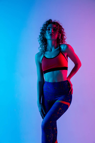 Portrait of young beautiful woman in bright sportwear isolated on gradient pink-blue studio background in neon light. - Foto, Imagen