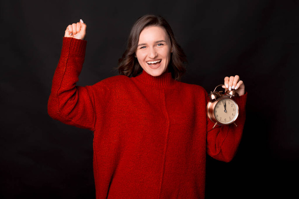 Aufgeregte, fröhliche junge Frau im roten Pullover hält Uhr bei Siegergeste auf dunklem Hintergrund - Foto, Bild