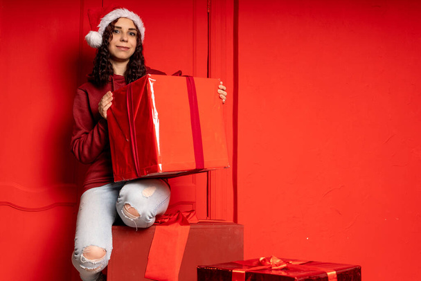 Young attractive woman in Santa Claus hat sits on gifts near red wall. Smiley female sitting on big box of gift. Concept of holidays, gifts and good mood - Foto, immagini