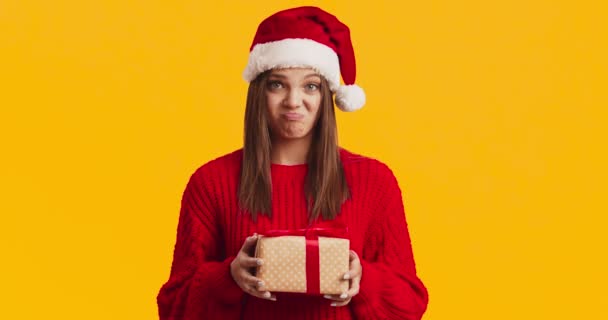 Młoda niezadowolona kobieta w kapeluszu Mikołaja trzyma pudełko prezentów i zmarszczki, pomarańczowe tło studio - Materiał filmowy, wideo