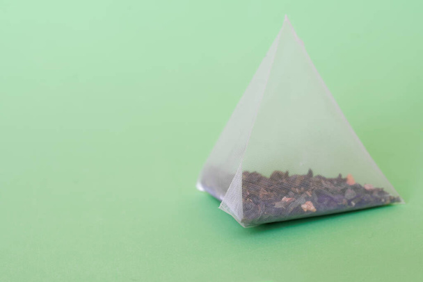 Una bolsa de té piramidal con té verde, hojas de menta y sabores de frutas se encuentra en una superficie verde. Espacio libre para una inscripción. Macro. Profundidad superficial del campo  - Foto, imagen