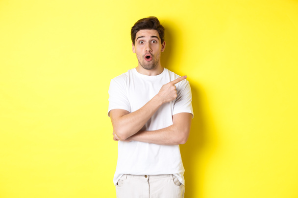 Portrét překvapeného pohledného muže ukazujícího prstem doprava, udýchaný, stojící nad žlutým pozadím - Fotografie, Obrázek