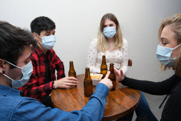 Vier Freunde mit Gesichtsmasken in einer Kneipe verbringen Zeit miteinander. Neue Normalität in Bars. - Foto, Bild
