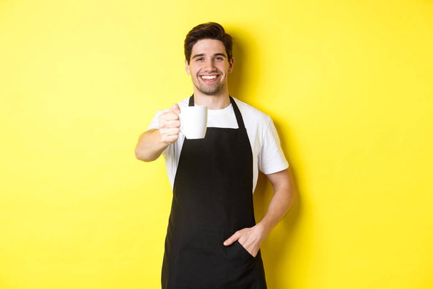 Knappe barista in zwart schort geven u kopje koffie en glimlachen, staande over gele achtergrond - Foto, afbeelding