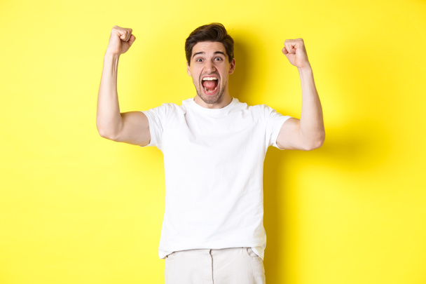 Obrázek vzrušeného muže, jak vyhrává, zvedá ruce a oslavuje, vítězí a fandí týmu, stojí nad žlutým pozadím - Fotografie, Obrázek