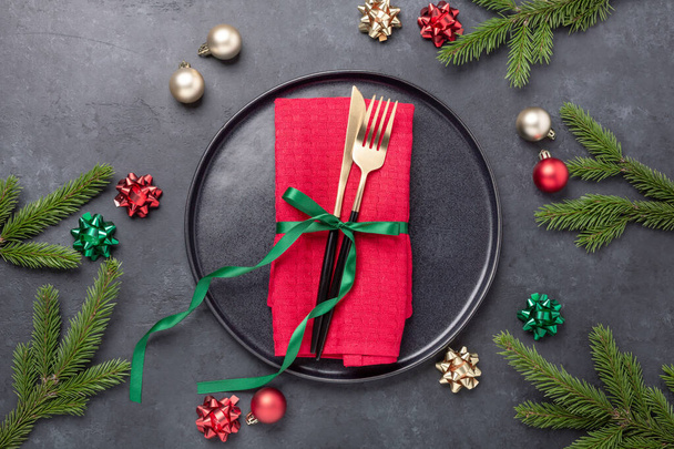 Christmas table setting with black ceramic plate, fir tree branch and gold and red accessories on black stone background. Top view. Copy space - Image - Zdjęcie, obraz