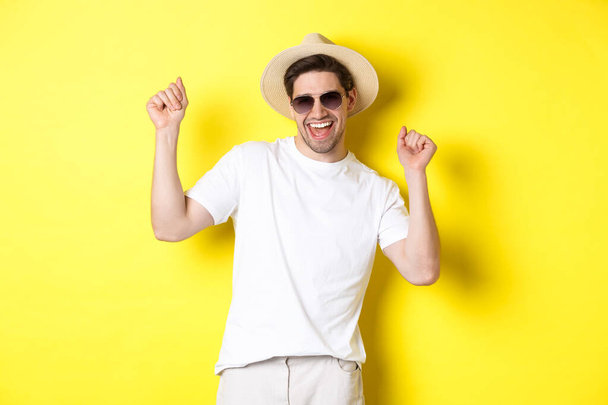 Tourism, travelling and holidays concept. Happy caucasian guy dancing and having fun on vacation, wearing sunglasses with straw hat, standing against yellow background - Photo, Image
