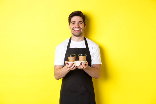 Amistoso barista en delantal negro dando orden de comida para llevar, sosteniendo dos tazas de café y sonriendo, de pie sobre fondo amarillo - Foto, Imagen