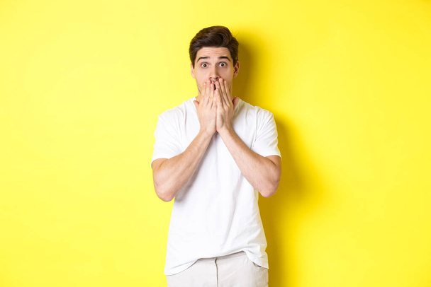 Handsome man looking shocked and speechless, holding hands on mouth, standing over yellow background - Fotoğraf, Görsel