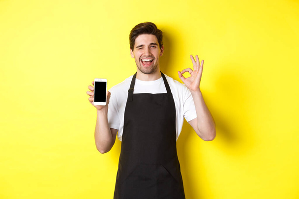 Schöne Coffeeshop-Mitarbeiterin zeigt ok Schild und Smartphone-Bildschirm, empfiehlt Anwendung, steht über gelbem Hintergrund - Foto, Bild