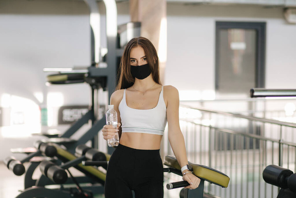 Attractive brunette female have a break in gym. Woman in protective mask and hold bottle of water - Photo, Image