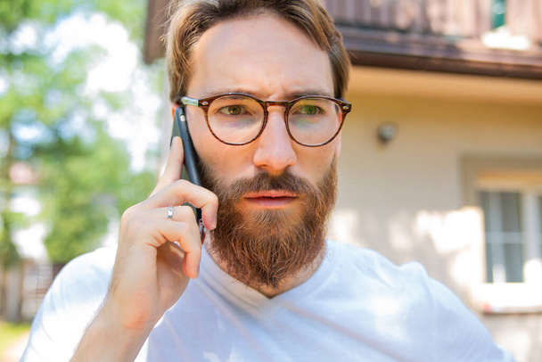 Receiving bad news via cell phone. Close-up of a young grim-faced man talking on a mobile phone while standing outdoor. - 写真・画像