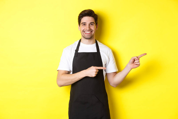 Ystävällinen tarjoilija osoittaa sormella oikealle, näyttää logon tai promo tarjous, yllään musta esiliina yhtenäinen, seisoo yli keltainen tausta - Valokuva, kuva