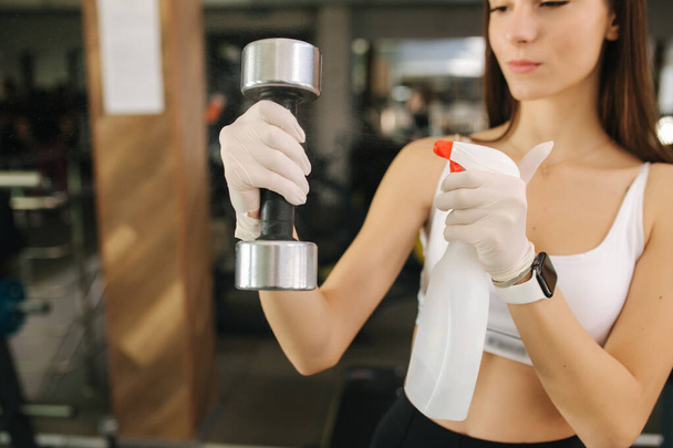 Young brunette woman offers a sanitizer to clean our hands and dembbells at the gym. Woman in white gloves - Photo, Image