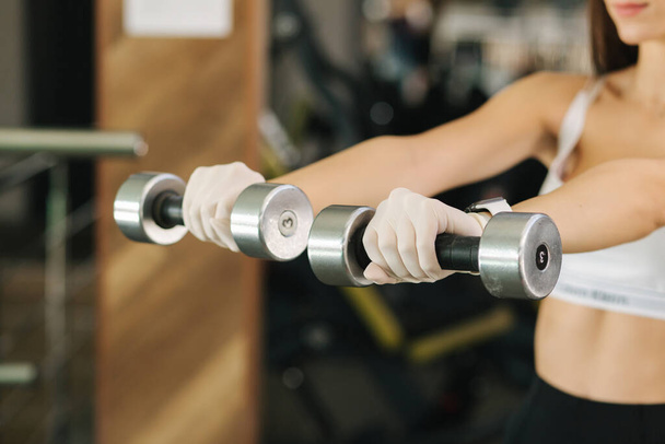 Brunette woman in white gloves hold two dumbbells in hand. Female training in the gym - Foto, imagen