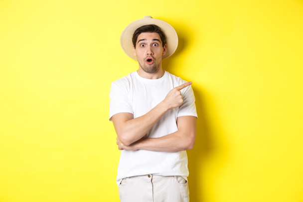 Surprised man in straw hat pointing finger right, showing promo banner, standing over yellow background - Photo, Image