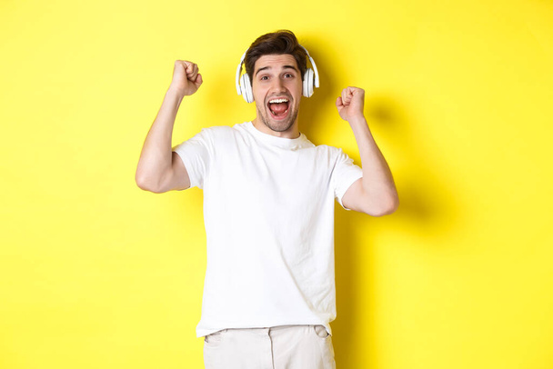 Opgewonden knappe man die danst en meezingt, muziek luistert in een koptelefoon, over een gele achtergrond staat - Foto, afbeelding