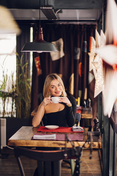 Smiling business woman in a restaurant holding a Cup of tea and looking at the camera, sitting near the window in a black dress - Foto, afbeelding