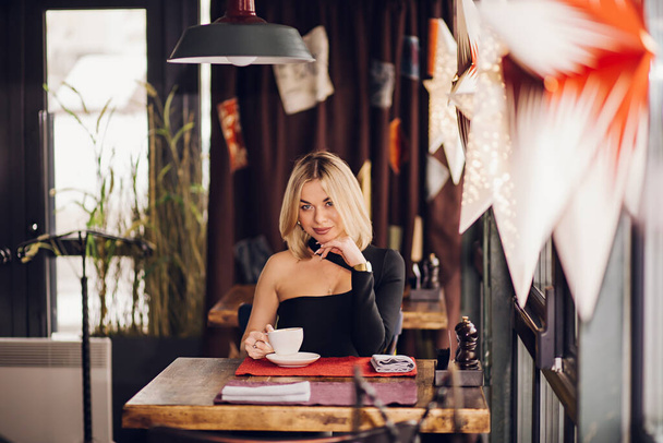 A successful confident business woman sits near the window in a cafe, restaurant. Blonde girl puts on a black dress - 写真・画像