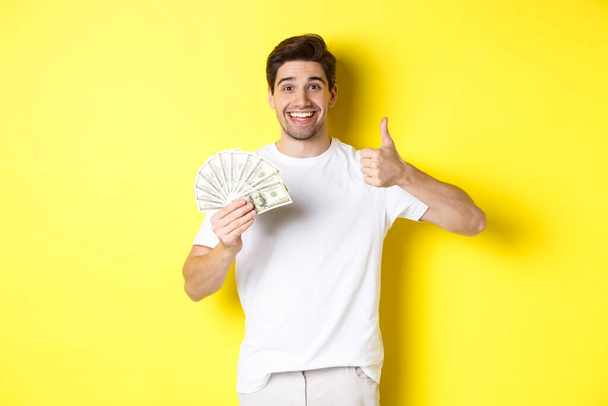 Ικανοποιημένος αγοραστής άνθρωπος δείχνει αντίχειρας-up και κρατώντας χρήματα, ψώνια με μετρητά, στέκεται πάνω από κίτρινο φόντο - Φωτογραφία, εικόνα