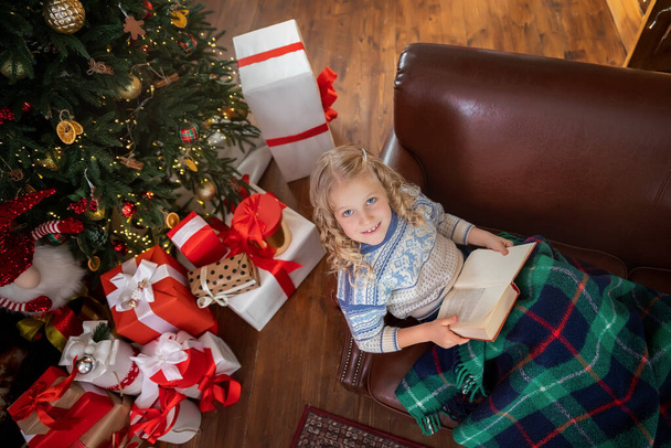 Cute adorable caucasian blond female kid reading fairytale story book sitting on sofa with warm plaid near decorated christmas tree with many gifts. Sweet liitle child at new year eve in cozy room - Photo, Image